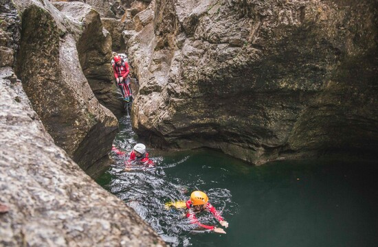 Personen beim Canyoning in einer felsigen Schlucht, ausgerüstet mit Helmen und Neoprenanzügen, schwimmen durch klares Wasser.