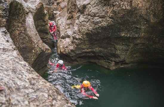 Personen beim Canyoning in einer felsigen Schlucht, ausgerüstet mit Helmen und Neoprenanzügen, schwimmen durch klares Wasser.