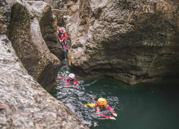 Personen beim Canyoning in einer felsigen Schlucht, ausgerüstet mit Helmen und Neoprenanzügen, schwimmen durch klares Wasser.